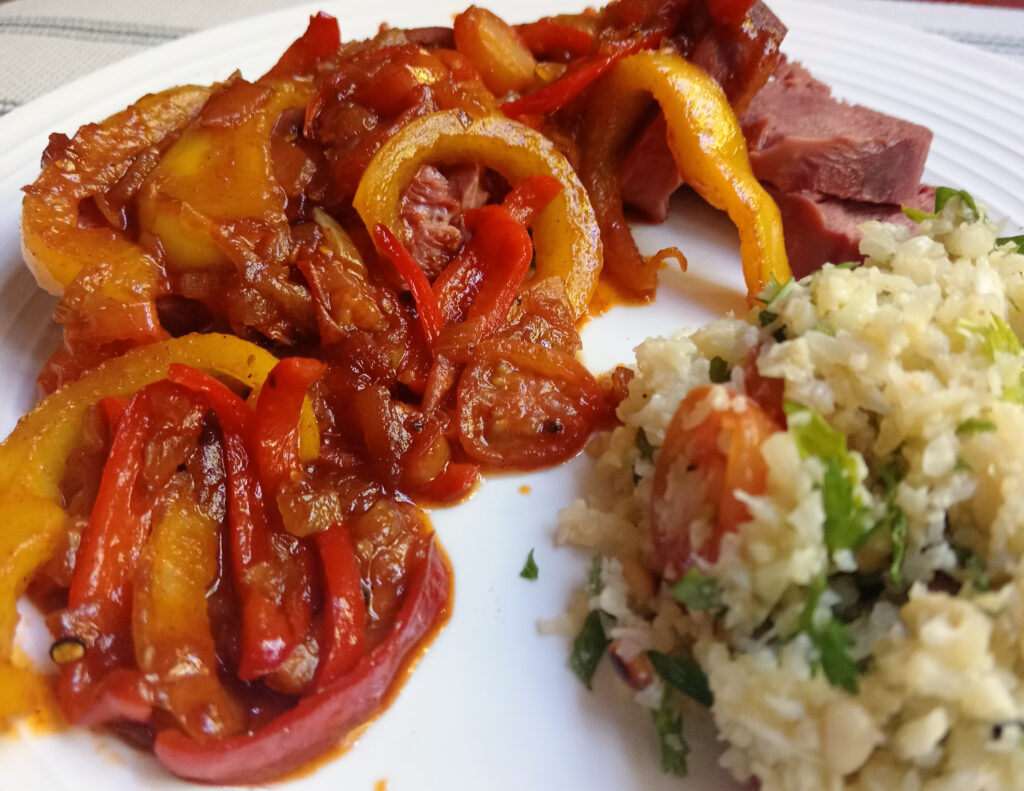 Close-up of sliced beef tongue topped with a glossy bell pepper chutney in shades of red and yellow, served on a white plate with a small portion of cauliflower salad.