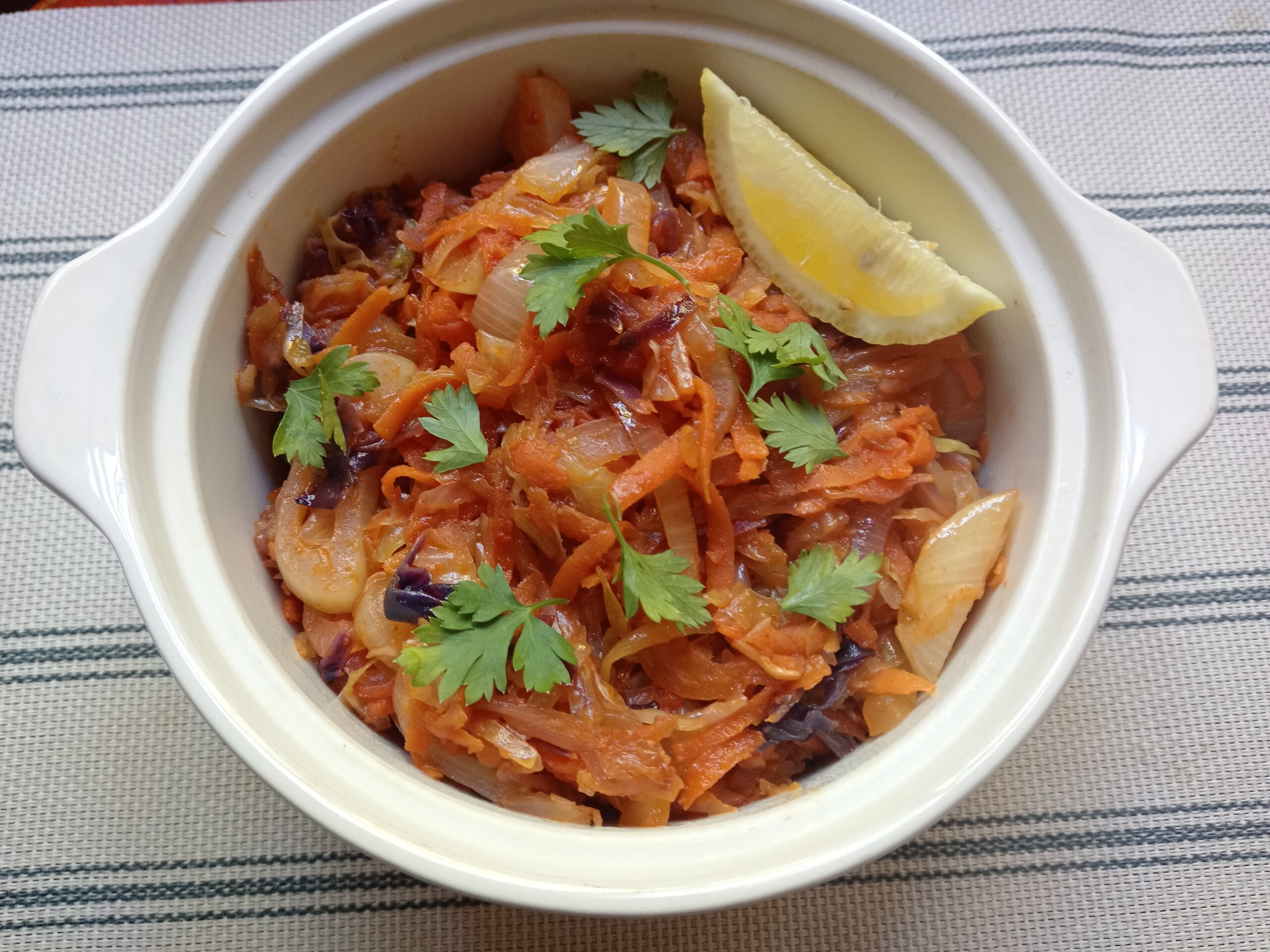 Braised cabbage with tomato in a cream-coloured casserole dish, garnished with fresh parsley and a lemon wedge on a striped cloth background.