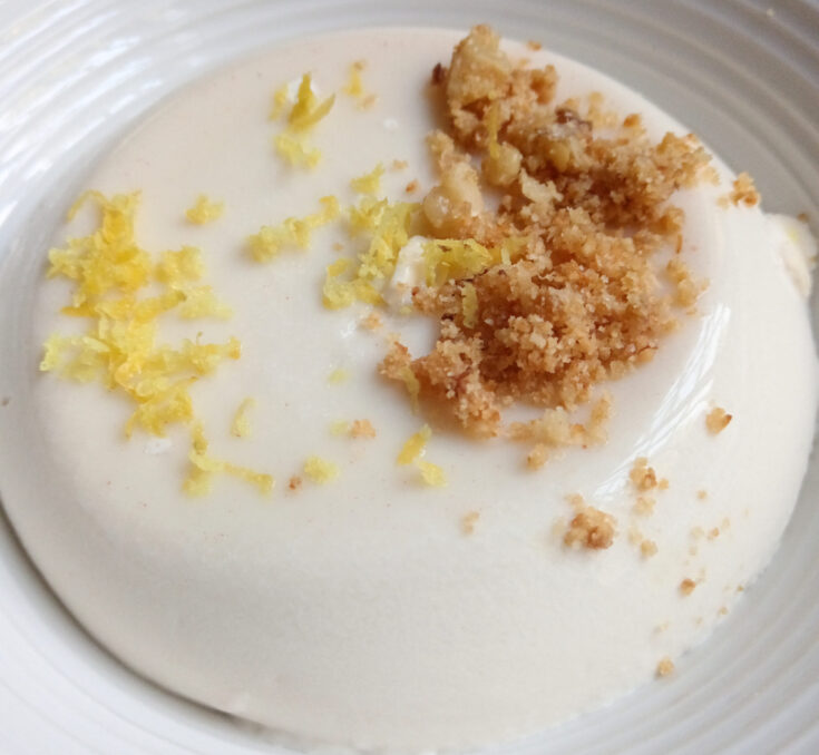 Buttermilk panna cotta topped with toasted almond and coconut crumb and fresh lemon zest on a white plate.