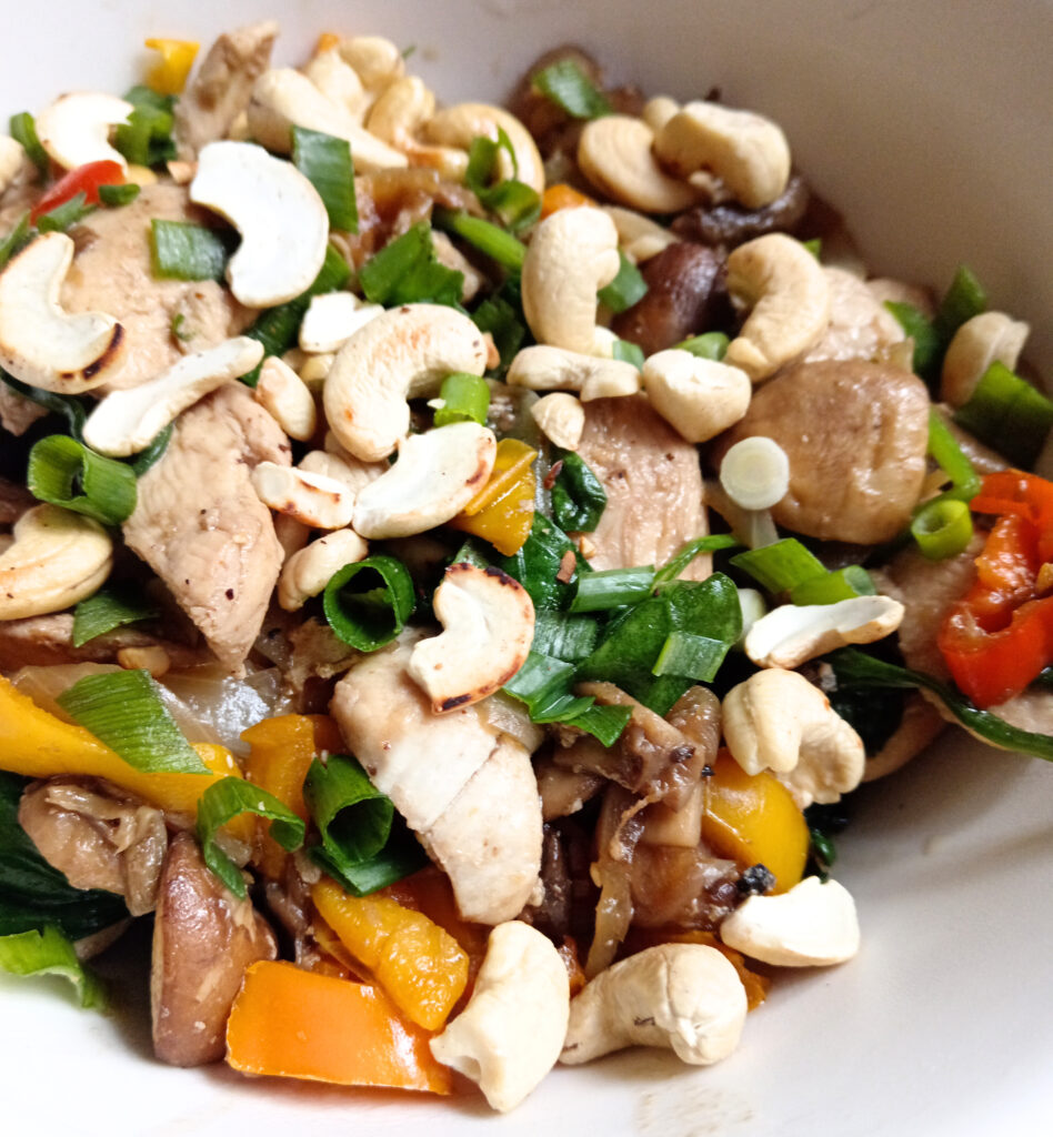 Keto cashew chicken stir-fry with mushrooms, sweet baby peppers, baby spinach, toasted cashews, and spring onions served in a white bowl.