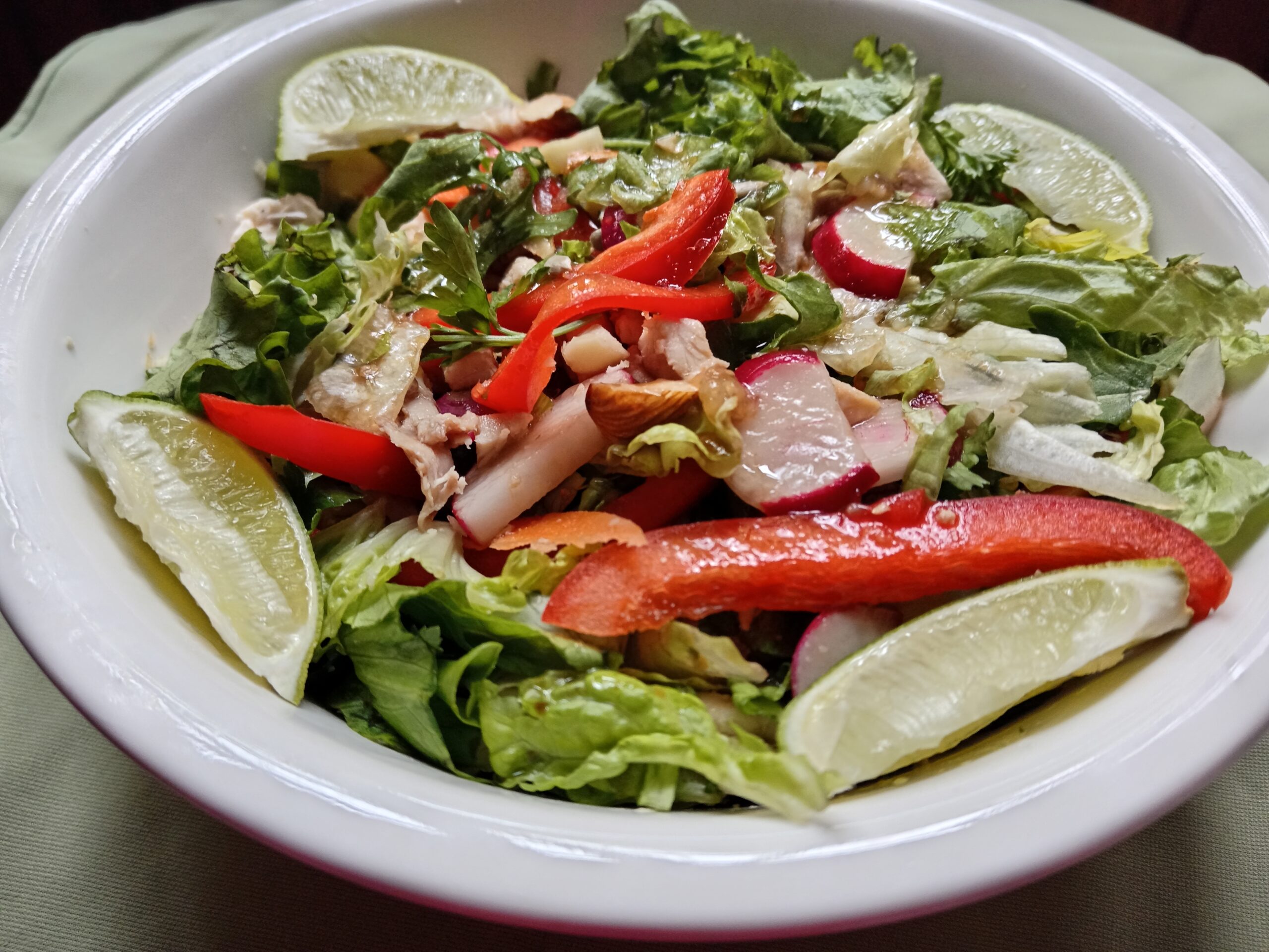 A vibrant chicken salad in a white bowl with red bell pepper, radish, fresh greens, and lime wedges, lightly coated in sesame dressing.