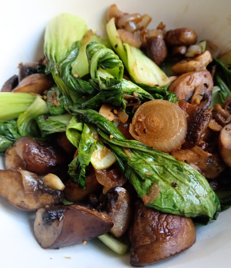 Butter-sautéed mushrooms, pak choi and shallots in a white serving bowl, finished with ginger, soy sauce, sesame oil and fresh lime.