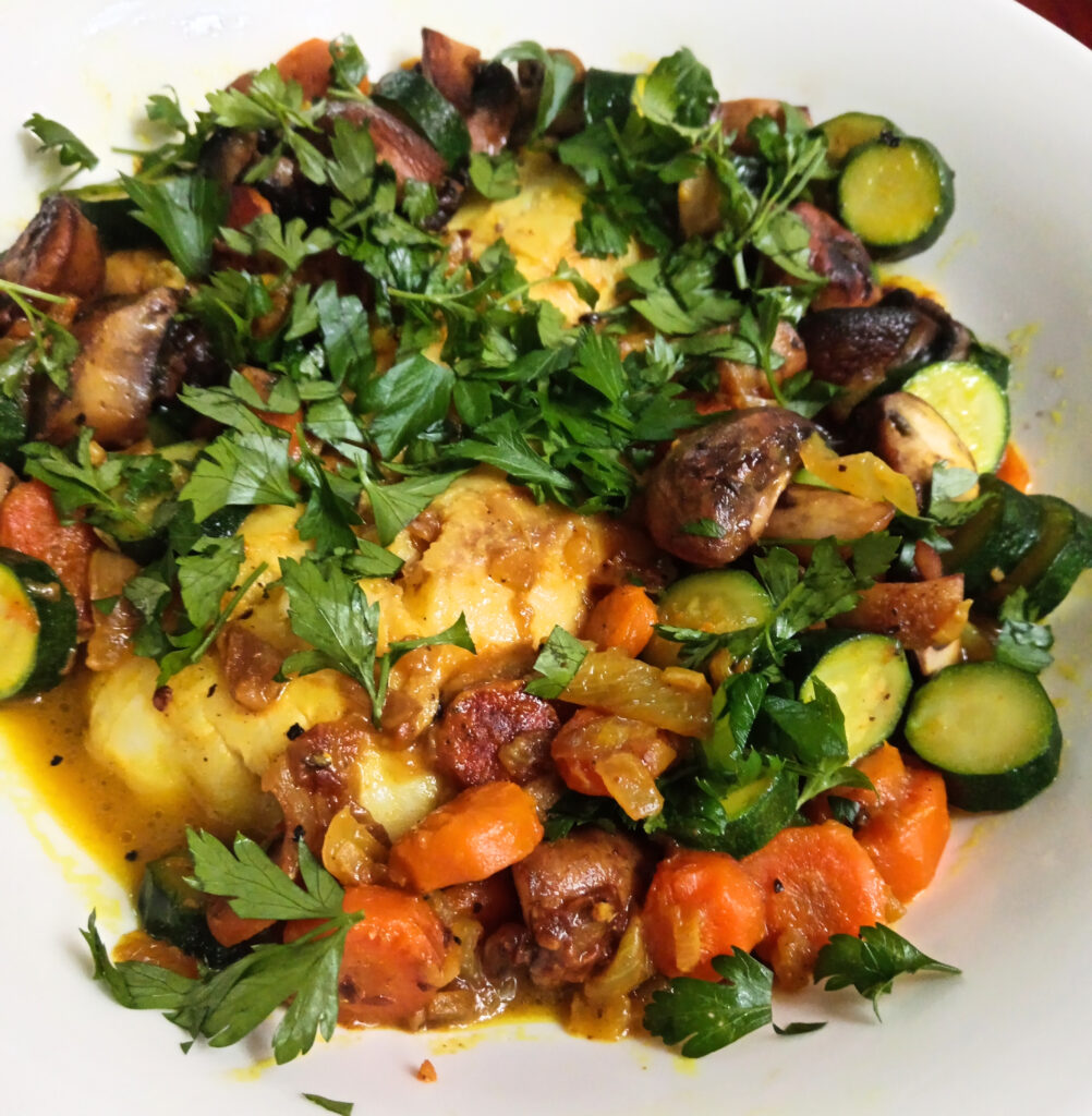 Butter-poached hake with turmeric, baby marrow, carrots, mushrooms, fresh parsley, and lime served in a shallow bowl.