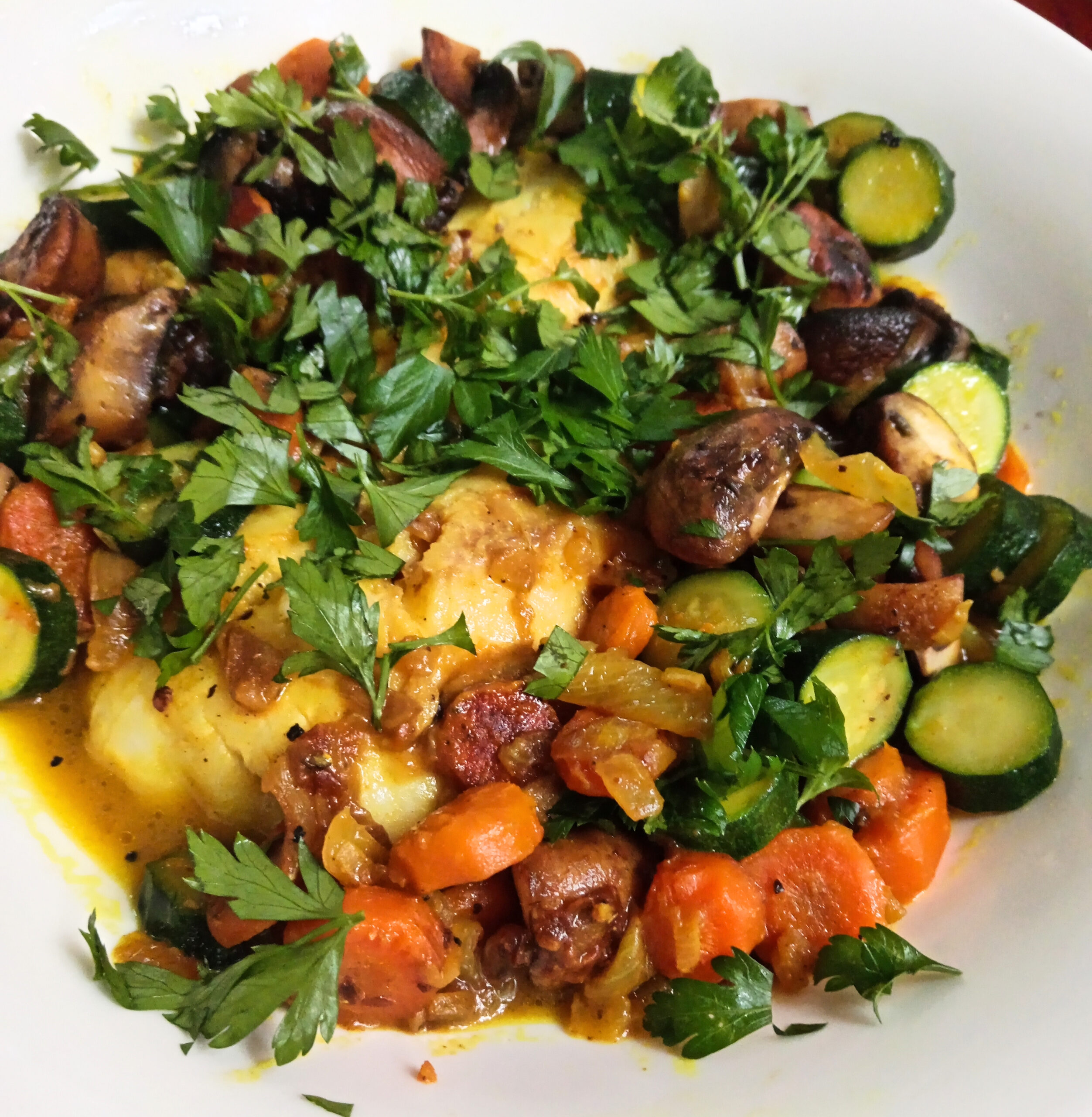 Butter-poached hake with turmeric, baby marrow, carrots, mushrooms, fresh parsley, and lime served in a shallow bowl.